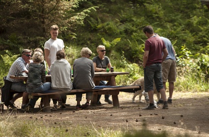 En lille forsamling mennesker er mødtes ude i skoven og sidder ved et borde-bænke-sæt