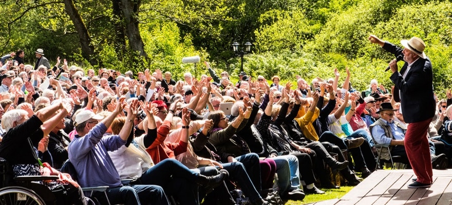 Hundredevis af ældre mennesker sidder til udendørs koncert med hænderne i vejret, med sol og skov i baggrunden
